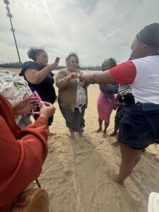 A small group of women gather together barefoot on a sandy beach under an overcast sky. In the center, a woman wearing a tan blazer and a black-and-white striped skirt ties a small string or bracelet onto another woman's wrist. On the left, another woman records the moment on her smartphone, while someone in the foreground holds small pink strips of paper. A tall cell tower structure stands far in the background.