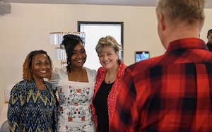 Three women stand side-by-side inside a room, smiling warmly as they pose for a photo. The woman on the left has shoulder-length brown hair and wears a blue patterned blouse. The woman in the center has styled black hair and wears a white dress with red and green floral embroidery. The woman on the right has blonde hair and wears a vibrant coral or pink blazer over a black top. In the foreground, the back of a man wearing a red and black plaid flannel shirt is visible as he faces them.