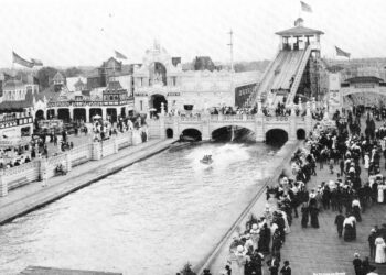 A vintage black-and-white photograph shows a packed crowd dressed in early 20th-century attire walking along a boardwalk and bridge at an amusement park. In the center, a long water lagoon features a flat-bottomed boat skipping across the surface after sliding down a massive "Shoot the Chutes" water ramp visible in the background. Ornate, white Beaux-Arts-style buildings with arches, towers, and flags line the waterfront under a clear sky