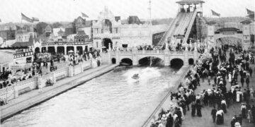 A vintage black-and-white photograph shows a packed crowd dressed in early 20th-century attire walking along a boardwalk and bridge at an amusement park. In the center, a long water lagoon features a flat-bottomed boat skipping across the surface after sliding down a massive "Shoot the Chutes" water ramp visible in the background. Ornate, white Beaux-Arts-style buildings with arches, towers, and flags line the waterfront under a clear sky