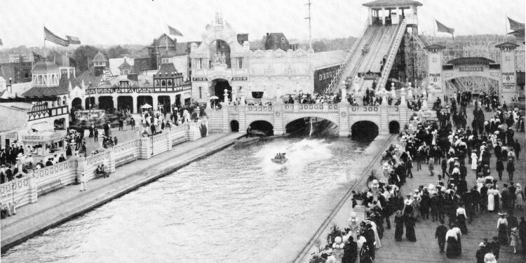 A vintage black-and-white photograph shows a packed crowd dressed in early 20th-century attire walking along a boardwalk and bridge at an amusement park. In the center, a long water lagoon features a flat-bottomed boat skipping across the surface after sliding down a massive "Shoot the Chutes" water ramp visible in the background. Ornate, white Beaux-Arts-style buildings with arches, towers, and flags line the waterfront under a clear sky