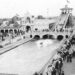 A vintage black-and-white photograph shows a packed crowd dressed in early 20th-century attire walking along a boardwalk and bridge at an amusement park. In the center, a long water lagoon features a flat-bottomed boat skipping across the surface after sliding down a massive "Shoot the Chutes" water ramp visible in the background. Ornate, white Beaux-Arts-style buildings with arches, towers, and flags line the waterfront under a clear sky