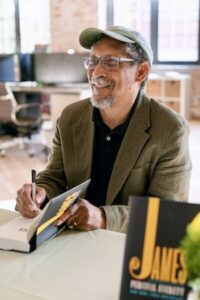 Candid indoor shot of author Percival Everett sitting at a table covered in a white cloth during a book signing event. He is looking slightly to the right with a warm smile, wearing round glasses, a light olive green baseball cap, a dark polo shirt, and a brown corduroy blazer. In his right hand, he holds a black pen, writing a personalized inscription inside an open hardcover book. In the foreground, another copy of his award-winning novel, James, is prominently displayed but softly out of focus, showcasing the bold yellow and black cover design. The background shows a brightly lit, modern room with industrial windows and office furniture.