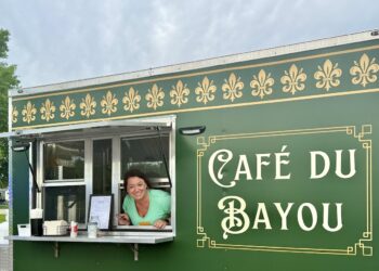 A cheerful woman in a light green top looks out from the open service window of a dark green food truck, leaning her arms against the silver counter ledge. The side of the vehicle features ornate gold border framing enclosing the words "Café du Bayou" in a stylized white font. A row of golden fleur-de-lis symbols lines the upper border of the truck paneling. A framed menu rests on the counter shelf to the left under a cloudy sky.