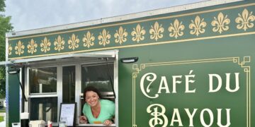 A cheerful woman in a light green top looks out from the open service window of a dark green food truck, leaning her arms against the silver counter ledge. The side of the vehicle features ornate gold border framing enclosing the words "Café du Bayou" in a stylized white font. A row of golden fleur-de-lis symbols lines the upper border of the truck paneling. A framed menu rests on the counter shelf to the left under a cloudy sky.