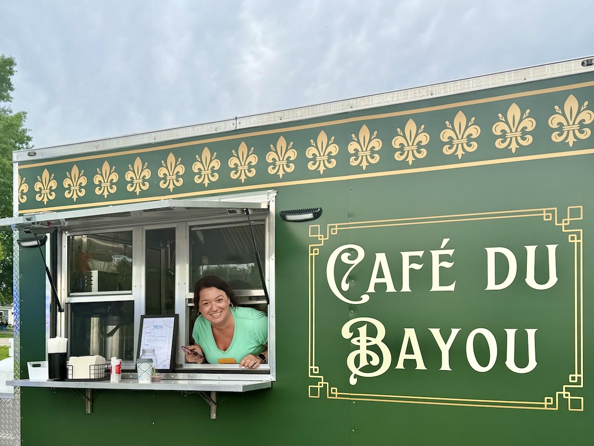 A cheerful woman in a light green top looks out from the open service window of a dark green food truck, leaning her arms against the silver counter ledge. The side of the vehicle features ornate gold border framing enclosing the words "Café du Bayou" in a stylized white font. A row of golden fleur-de-lis symbols lines the upper border of the truck paneling. A framed menu rests on the counter shelf to the left under a cloudy sky.