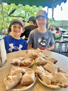 Two smiling boys sit side-by-side at a round table on an outdoor patio under a green awning. The younger boy on the left wears a blue Under Armour athletic shirt and a tan cap. The older boy on the right wears a grey shirt featuring Mickey Mouse wearing sunglasses and a grey cap. On the table in front of them are four small plates heavily stacked with golden-brown, square-shaped beignets buried under a thick coating of white powdered sugar. The blurry background shows other diners sitting at patio tables near trees and a parking lot.