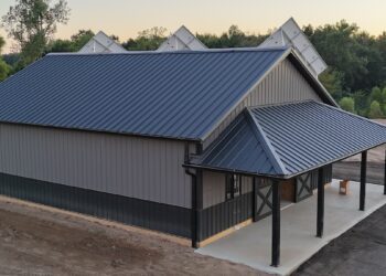 High-angle shot of a newly constructed pole barn with a dark grey metal roof and matching vertical siding. The building features a lower dark wainscoting panel, a covered concrete porch with supportive black pillars, and barn doors. Solar panels are visible peaking out from behind the main roofline.