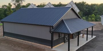 High-angle shot of a newly constructed pole barn with a dark grey metal roof and matching vertical siding. The building features a lower dark wainscoting panel, a covered concrete porch with supportive black pillars, and barn doors. Solar panels are visible peaking out from behind the main roofline.