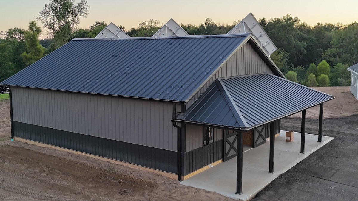 High-angle shot of a newly constructed pole barn with a dark grey metal roof and matching vertical siding. The building features a lower dark wainscoting panel, a covered concrete porch with supportive black pillars, and barn doors. Solar panels are visible peaking out from behind the main roofline.