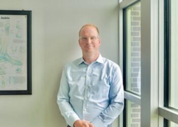 Three-quarter length indoor portrait of a medical provider leaning against a white wall next to a large multi-paned window. The man has short reddish-blonde hair, wears glasses, a light blue button-down dress shirt, and grey trousers with his hands clasped loosely in front of him. To his right, a framed medical illustration chart titled "Ankle" hangs on the wall, showing various diagrams of foot anatomy. Soft, natural morning light streams in through the window.