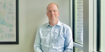 Three-quarter length indoor portrait of a medical provider leaning against a white wall next to a large multi-paned window. The man has short reddish-blonde hair, wears glasses, a light blue button-down dress shirt, and grey trousers with his hands clasped loosely in front of him. To his right, a framed medical illustration chart titled "Ankle" hangs on the wall, showing various diagrams of foot anatomy. Soft, natural morning light streams in through the window.