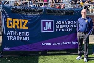 Full-length outdoor shot of a man standing on an athletic field turf next to a large promotional banner structure. The man is wearing glasses, a navy blue long-sleeve polo shirt with "Griz Athletics" branding on the chest, dark grey trousers, and white athletic sneakers. The large navy banner next to him displays "GRIZ ATHLETIC TRAINING" in bold yellow and white lettering on the left. The right side features the purple and white logo for "Johnson Memorial Health" alongside their slogan, "Great care starts here." In the background, crowds of spectators are visible sitting in outdoor stadium bleachers.