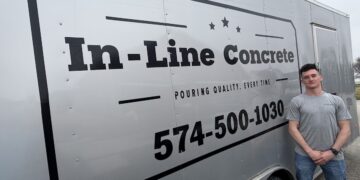 A man in a grey t-shirt and jeans stands next to a large silver commercial trailer. The trailer features a prominent black logo reading "In-Line Concrete" with the slogan "POURING QUALITY, EVERY TIME" and the phone number 574-500-1030 printed in large black font.