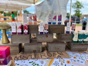 Eye-level shot of a product display at a farmers market booth. Small, rectangular product boxes feature minimalist branding with distinct botanical illustrations in shades of pink, orange, purple, and green. They are artfully stacked on and inside dark, weathered wooden crates of varying sizes. To the far left, a decorative brass mushroom figurine stands on the table. The display sits on a patterned floral tablecloth over a strip of natural burlap cloth stenciled with the word "AMSTERDAM" in bold black lettering. The background is softly blurred, showing glimpses of outdoor market tents.