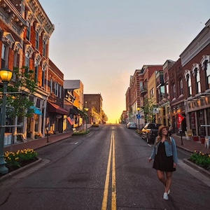 A wide, low-angle outdoor photograph shows Main Street in historic downtown Franklin, Tennessee during a vibrant golden hour sunset. A woman in a black dress and denim jacket walks along the asphalt street next to double yellow lines. Classic multi-story brick buildings with historic storefronts, awnings, and lit street lamps line both sides of the road, leading up a slight hill where the bright sun dips below the horizon.