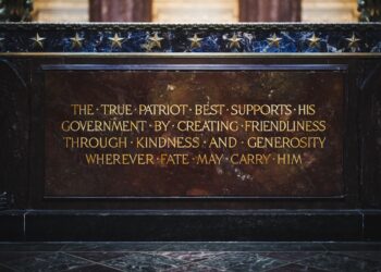 A close-up shot of an inscribed inscription plaque mounted on a dark marble or granite structure, identified as part of the Indiana World War Memorial. The plaque is a rectangular slab of polished reddish-brown marble with gilded capital lettering that reads: "THE TRUE PATRIOT BEST SUPPORTS HIS GOVERNMENT BY CREATING FRIENDLINESS THROUGH KINDNESS AND GENEROSITY WHEREVER FATE MAY CARRY HIM." Bullet points separate the first few words of the inscription. Above the plaque, a horizontal band of dark, veined blue marble is adorned with a row of small, metallic gold stars.