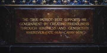 A close-up shot of an inscribed inscription plaque mounted on a dark marble or granite structure, identified as part of the Indiana World War Memorial. The plaque is a rectangular slab of polished reddish-brown marble with gilded capital lettering that reads: "THE TRUE PATRIOT BEST SUPPORTS HIS GOVERNMENT BY CREATING FRIENDLINESS THROUGH KINDNESS AND GENEROSITY WHEREVER FATE MAY CARRY HIM." Bullet points separate the first few words of the inscription. Above the plaque, a horizontal band of dark, veined blue marble is adorned with a row of small, metallic gold stars.