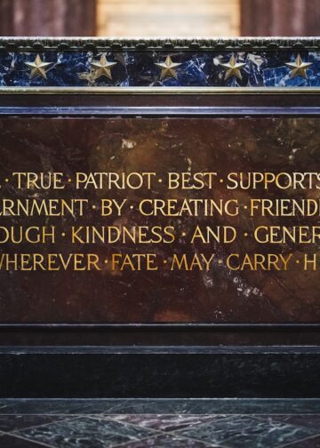 A close-up shot of an inscribed inscription plaque mounted on a dark marble or granite structure, identified as part of the Indiana World War Memorial. The plaque is a rectangular slab of polished reddish-brown marble with gilded capital lettering that reads: "THE TRUE PATRIOT BEST SUPPORTS HIS GOVERNMENT BY CREATING FRIENDLINESS THROUGH KINDNESS AND GENEROSITY WHEREVER FATE MAY CARRY HIM." Bullet points separate the first few words of the inscription. Above the plaque, a horizontal band of dark, veined blue marble is adorned with a row of small, metallic gold stars.