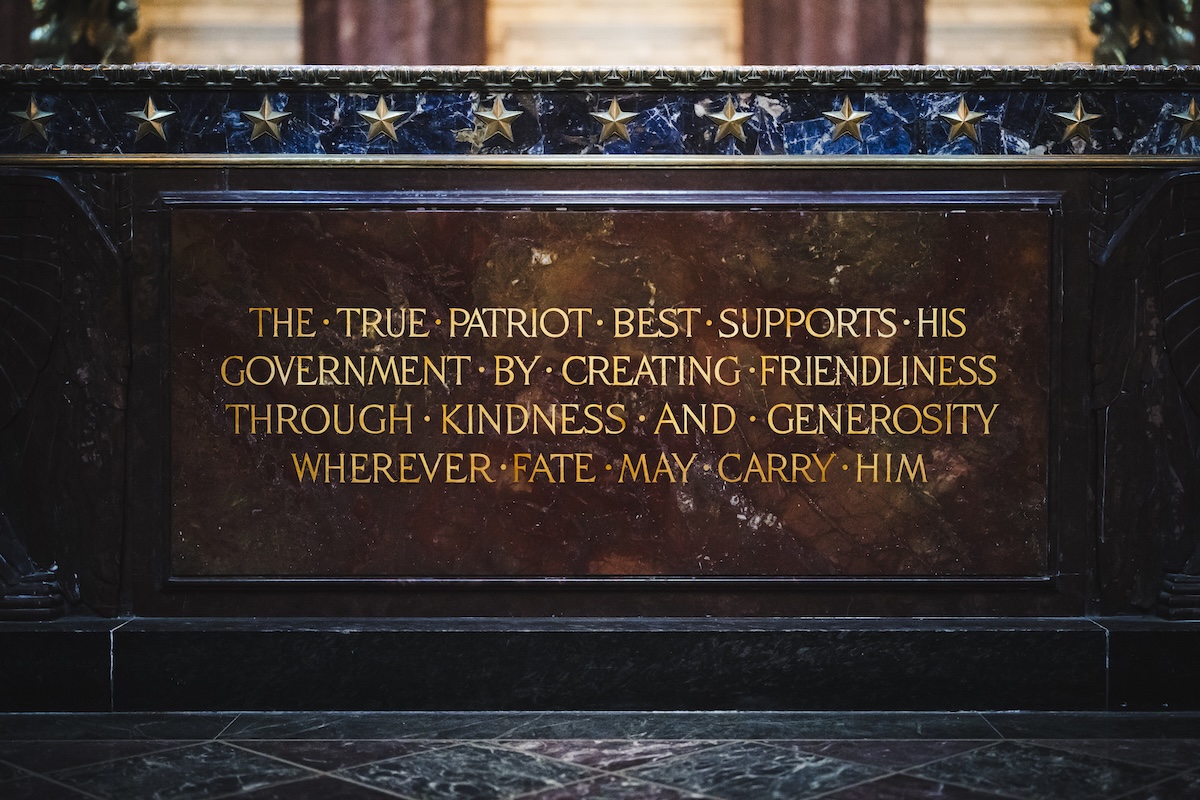 A close-up shot of an inscribed inscription plaque mounted on a dark marble or granite structure, identified as part of the Indiana World War Memorial. The plaque is a rectangular slab of polished reddish-brown marble with gilded capital lettering that reads: "THE TRUE PATRIOT BEST SUPPORTS HIS GOVERNMENT BY CREATING FRIENDLINESS THROUGH KINDNESS AND GENEROSITY WHEREVER FATE MAY CARRY HIM." Bullet points separate the first few words of the inscription. Above the plaque, a horizontal band of dark, veined blue marble is adorned with a row of small, metallic gold stars.