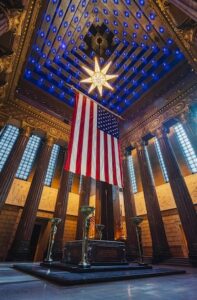 A low-angle, vertical shot capturing the grand and solemn interior of the Shrine Room inside the Indiana War Memorial Museum in Indianapolis. In the upper center, an enormous American flag hangs vertically from invisible wires. Suspended directly above the flag is the Star of Destiny, a prominent, illuminated Swedish crystal chandelier shaped like an eight-pointed star. The vast, 110-foot-tall ceiling rises in recessed tiers, painted deep blue and embedded with rows of tiny, twinkling blue lights resembling stars. Surrounding the room are towering, 40-foot fluted columns crafted from dark red Vermont marble. In the lower center, beneath the flag, sits the dark stone Altar of Consecration on a tiered black marble platform, flanked by tall decorative torchieres. Tall, narrow stained-glass windows line the walls, casting a soft blue glow.