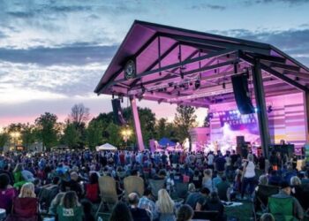 A large outdoor crowd sits in lawn chairs facing a modern open-air pavilion stage at the Nickel Plate District Amphitheater in Fishers, Indiana during an evening concert. The steel-framed wooden pavilion structure is illuminated from inside with vibrant pink and violet stage lights, casting a glow onto the musical artist performing live on stage. A backdrop screen on stage displays the band name "Manchester Orchestra" in white block letters. The audience fills the green grass lawn stretching into the background under a twilight sky with soft, orange-tinted clouds.