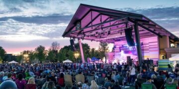 A large outdoor crowd sits in lawn chairs facing a modern open-air pavilion stage at the Nickel Plate District Amphitheater in Fishers, Indiana during an evening concert. The steel-framed wooden pavilion structure is illuminated from inside with vibrant pink and violet stage lights, casting a glow onto the musical artist performing live on stage. A backdrop screen on stage displays the band name "Manchester Orchestra" in white block letters. The audience fills the green grass lawn stretching into the background under a twilight sky with soft, orange-tinted clouds.