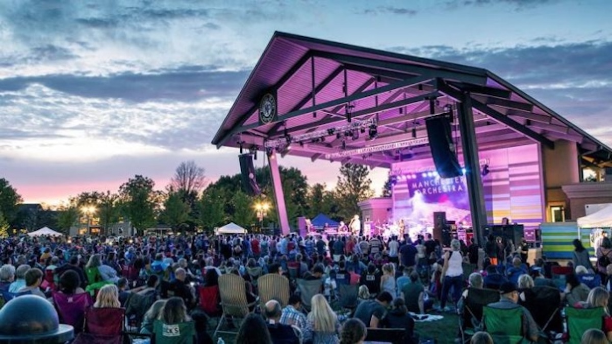 A large outdoor crowd sits in lawn chairs facing a modern open-air pavilion stage at the Nickel Plate District Amphitheater in Fishers, Indiana during an evening concert. The steel-framed wooden pavilion structure is illuminated from inside with vibrant pink and violet stage lights, casting a glow onto the musical artist performing live on stage. A backdrop screen on stage displays the band name "Manchester Orchestra" in white block letters. The audience fills the green grass lawn stretching into the background under a twilight sky with soft, orange-tinted clouds.