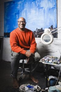 A full-length portrait of a smiling Black man with a shaved head and eyeglasses, sitting barefoot on a rolling stool in an art studio. He wears a bright orange sweatshirt and grey sweatpants. He sits directly in front of a very large canvas displaying a textured blue painting with hints of a crowd scene along the bottom edge. To his right, a side table holds an artist's palette covered in various paint colors, alongside tubes of paint and an illuminated magnifying glass lamp. The studio floor around him shows paint stains and used cloths.