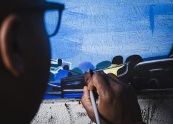 A close-up, over-the-shoulder view of an artist working on a canvas. A person with dark skin holds a paintbrush, applying paint to a detailed section of the artwork. The canvas displays a predominantly blue background with sketched outlines and painted sections depicting stylized figures and objects. Only the side of the artist's head and eyeglasses are visible in the foreground, out of focus.