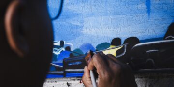 A close-up, over-the-shoulder view of an artist working on a canvas. A person with dark skin holds a paintbrush, applying paint to a detailed section of the artwork. The canvas displays a predominantly blue background with sketched outlines and painted sections depicting stylized figures and objects. Only the side of the artist's head and eyeglasses are visible in the foreground, out of focus.