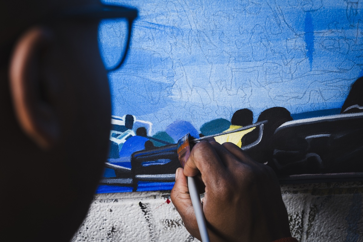 A close-up, over-the-shoulder view of an artist working on a canvas. A person with dark skin holds a paintbrush, applying paint to a detailed section of the artwork. The canvas displays a predominantly blue background with sketched outlines and painted sections depicting stylized figures and objects. Only the side of the artist's head and eyeglasses are visible in the foreground, out of focus.