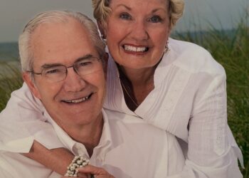 A close-up portrait of an older man and woman smiling warmly together outdoors. The woman, with short blonde hair, leans over the man's shoulder from behind, wrapping her arms around him and holding his hand. Both are dressed in matching white collared shirts. The man wears glasses, and the woman wears earrings, a necklace, a pearl bracelet, and a gold watch. The background shows a soft-focus setting of green beach grass dunes under an overcast sky.