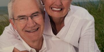 A close-up portrait of an older man and woman smiling warmly together outdoors. The woman, with short blonde hair, leans over the man's shoulder from behind, wrapping her arms around him and holding his hand. Both are dressed in matching white collared shirts. The man wears glasses, and the woman wears earrings, a necklace, a pearl bracelet, and a gold watch. The background shows a soft-focus setting of green beach grass dunes under an overcast sky.