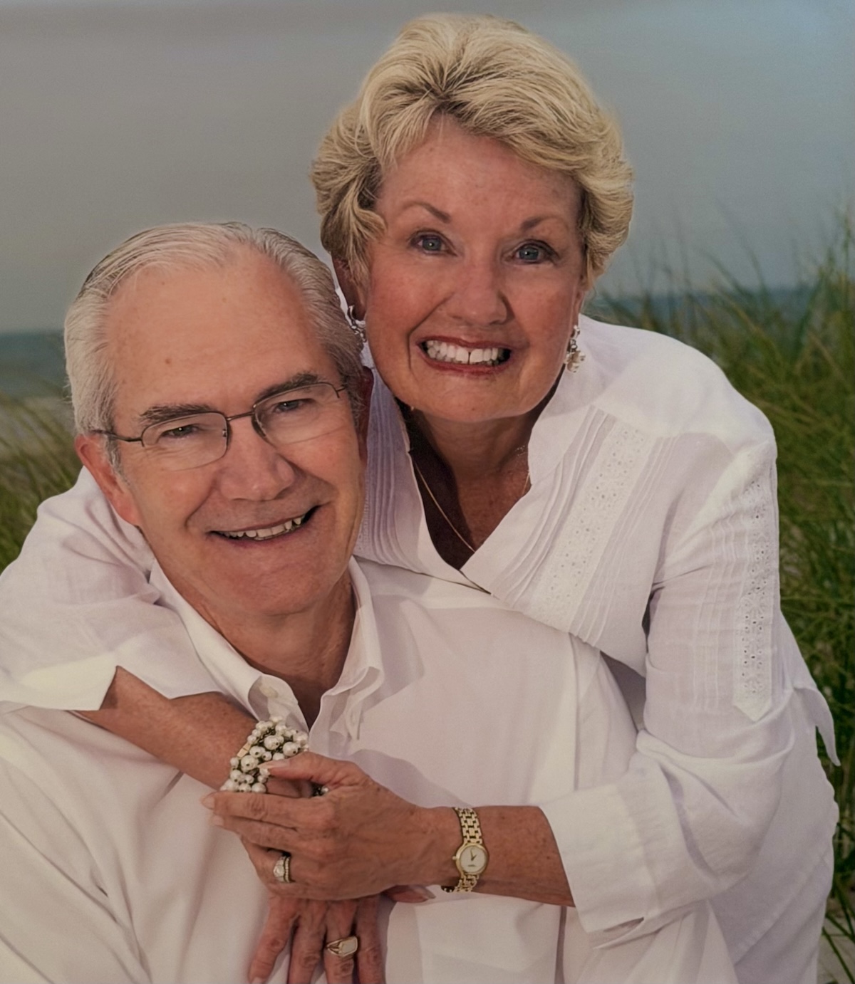 A close-up portrait of an older man and woman smiling warmly together outdoors. The woman, with short blonde hair, leans over the man's shoulder from behind, wrapping her arms around him and holding his hand. Both are dressed in matching white collared shirts. The man wears glasses, and the woman wears earrings, a necklace, a pearl bracelet, and a gold watch. The background shows a soft-focus setting of green beach grass dunes under an overcast sky.