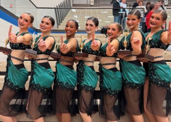 Seven smiling teenage female dancers stand in a line on wooden bleachers inside a gymnasium, celebrating a victory. They wear matching performance costumes featuring dark emerald green satin fabric draped over black and sheer mesh bodices embellished with sequins. Each dancer holds a gold trophy with a wooden base in one hand while raising the index finger of their other hand to signal "number one." Spectators are visible in the background seating.