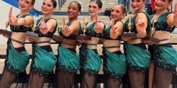 Seven smiling teenage female dancers stand in a line on wooden bleachers inside a gymnasium, celebrating a victory. They wear matching performance costumes featuring dark emerald green satin fabric draped over black and sheer mesh bodices embellished with sequins. Each dancer holds a gold trophy with a wooden base in one hand while raising the index finger of their other hand to signal "number one." Spectators are visible in the background seating.