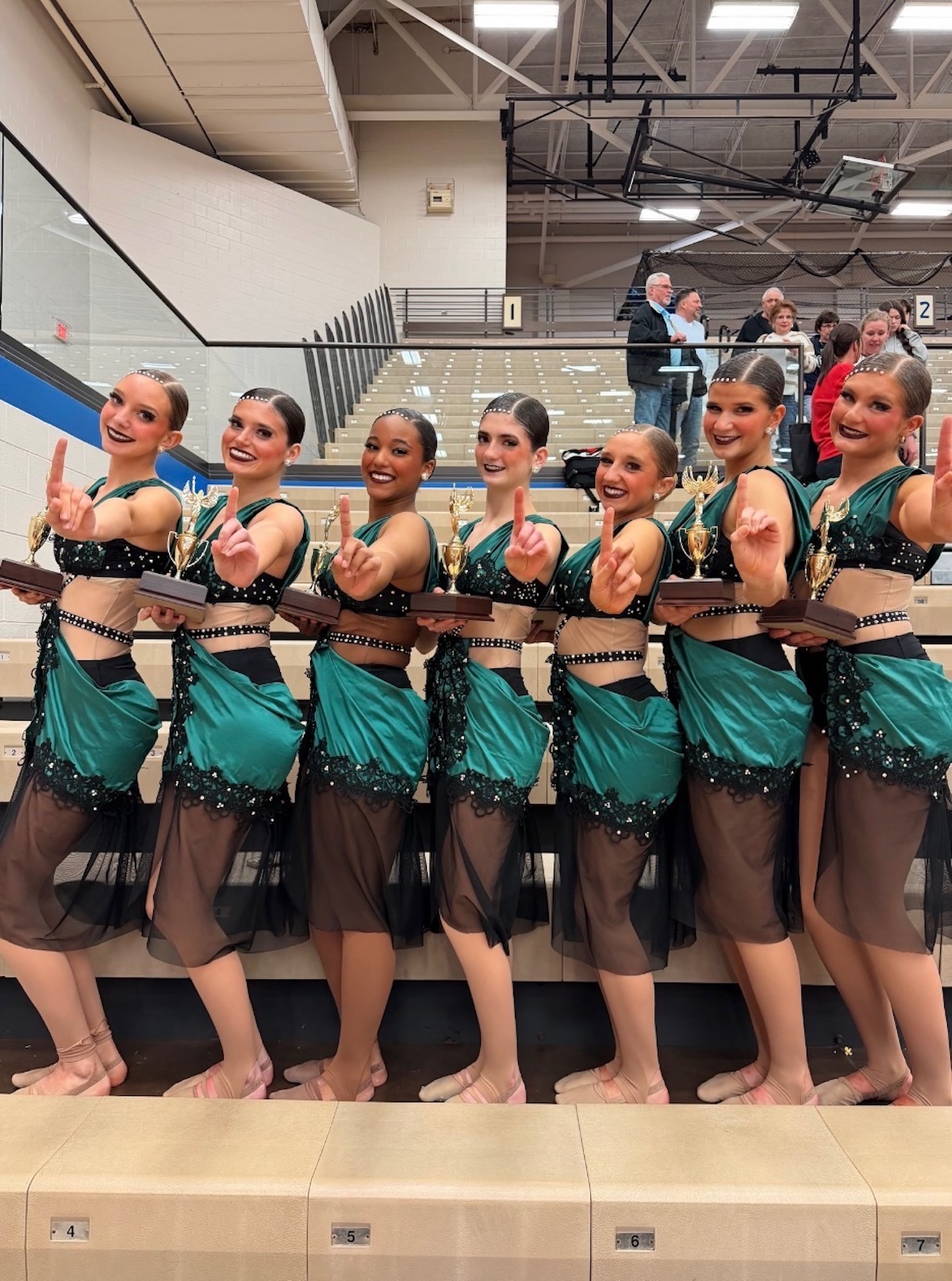 Seven smiling teenage female dancers stand in a line on wooden bleachers inside a gymnasium, celebrating a victory. They wear matching performance costumes featuring dark emerald green satin fabric draped over black and sheer mesh bodices embellished with sequins. Each dancer holds a gold trophy with a wooden base in one hand while raising the index finger of their other hand to signal "number one." Spectators are visible in the background seating.