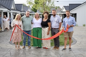 A group of six people stand smiling together on a paved stone courtyard for a ribbon-cutting ceremony. They hold a long, wide orange ribbon across the front of the group. Small white retail cottages with dark metal roofs frame the background space. A woman in a floral print poncho stands near an open cottage doorway on the far left.
