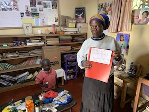 A woman wearing a white sweater, dark skirt, and a colorful blue and yellow headwrap stands inside a room holding open a red folder to display a white document. Next to her, a young child in a pink shirt stands behind a small table cluttered with various bottles and objects. The background features crowded wooden bookshelves packed with books, papers, a bulletin board, and educational posters.