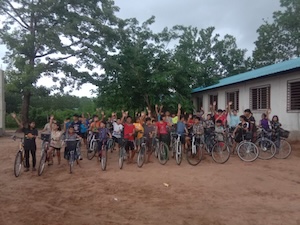 A large group of children and teenagers sit on bicycles in a dirt yard, cheering with their arms raised in the air. They are gathered outside a long, white, single-story building with a blue metal roof and barred windows. Lush green trees fill the background under a cloudy, overcast sky.