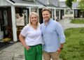 A smiling man and woman stand close together outside on a concrete walkway in front of a row of small, white commercial cottages with dark trim and dark metal roofs. The woman has blonde hair and wears a white V-neck t-shirt with emerald green wide-leg pants. The man has short blonde hair and wears a light blue button-down shirt with khaki shorts. Behind them, a sign on an easel features a photo of a person receiving a spa treatment.