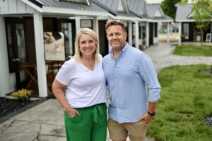 A smiling man and woman stand close together outside on a concrete walkway in front of a row of small, white commercial cottages with dark trim and dark metal roofs. The woman has blonde hair and wears a white V-neck t-shirt with emerald green wide-leg pants. The man has short blonde hair and wears a light blue button-down shirt with khaki shorts. Behind them, a sign on an easel features a photo of a person receiving a spa treatment.