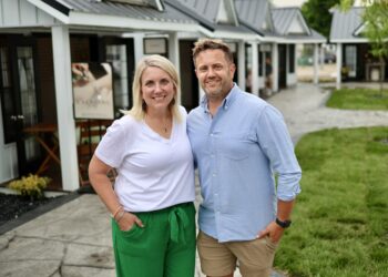 A smiling man and woman stand close together outside on a concrete walkway in front of a row of small, white commercial cottages with dark trim and dark metal roofs. The woman has blonde hair and wears a white V-neck t-shirt with emerald green wide-leg pants. The man has short blonde hair and wears a light blue button-down shirt with khaki shorts. Behind them, a sign on an easel features a photo of a person receiving a spa treatment.
