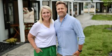 A smiling man and woman stand close together outside on a concrete walkway in front of a row of small, white commercial cottages with dark trim and dark metal roofs. The woman has blonde hair and wears a white V-neck t-shirt with emerald green wide-leg pants. The man has short blonde hair and wears a light blue button-down shirt with khaki shorts. Behind them, a sign on an easel features a photo of a person receiving a spa treatment.