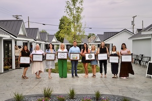 A line of ten business owners stand side-by-side in a paved outdoor courtyard between two rows of small white retail cottages. Each person holds a large framed certificate or business logo sign in front of them. A small green lawn area and a young tree sit behind the group under a hazy sky.