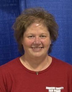 Studio headshot of a middle-aged woman smiling warmly at the camera. She has wavy, short brown hair and is wearing a maroon or dark red t-shirt. White text printed on the lower left of her shirt reads "JOHNSON COUNTY FAIR BOARD". She wears a thin metallic necklace with a small decorative charm. The background is a textured, solid blue curtain or fabric drape.