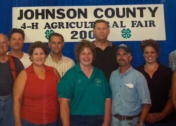 Flash photography shot of a group of nine people posing closely together inside a fairgrounds building. They stand in front of a large blue backdrop featuring a white sign with black text that reads "JOHNSON COUNTY 4-H AGRICULTURAL FAIR 2005" with two green four-leaf 4-H clovers. The individuals are dressed in casual summer clothing, including polo shirts, sleeveless tops, and jeans. One man on the far left wears distinctive orange suspenders over a dark t-shirt and blue jeans. Everyone is smiling directly at the camera.