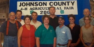 Flash photography shot of a group of nine people posing closely together inside a fairgrounds building. They stand in front of a large blue backdrop featuring a white sign with black text that reads "JOHNSON COUNTY 4-H AGRICULTURAL FAIR 2005" with two green four-leaf 4-H clovers. The individuals are dressed in casual summer clothing, including polo shirts, sleeveless tops, and jeans. One man on the far left wears distinctive orange suspenders over a dark t-shirt and blue jeans. Everyone is smiling directly at the camera.
