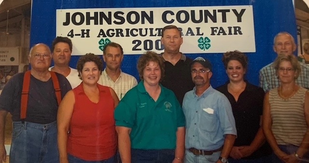Flash photography shot of a group of nine people posing closely together inside a fairgrounds building. They stand in front of a large blue backdrop featuring a white sign with black text that reads "JOHNSON COUNTY 4-H AGRICULTURAL FAIR 2005" with two green four-leaf 4-H clovers. The individuals are dressed in casual summer clothing, including polo shirts, sleeveless tops, and jeans. One man on the far left wears distinctive orange suspenders over a dark t-shirt and blue jeans. Everyone is smiling directly at the camera.